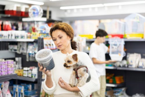 woman with curious Yorkshire terrier in hands engrossed in choosing dry dog food in pet store, attentively examining nutritional information on packaging